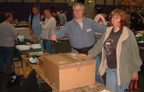 Photograph of Leicester 2009 FISTS stand showing Tony G3ZRJ, his XYL Sally and a large transceiver-size cardboard box.  In the background is John M0CDL and possibly Rob M0BPT